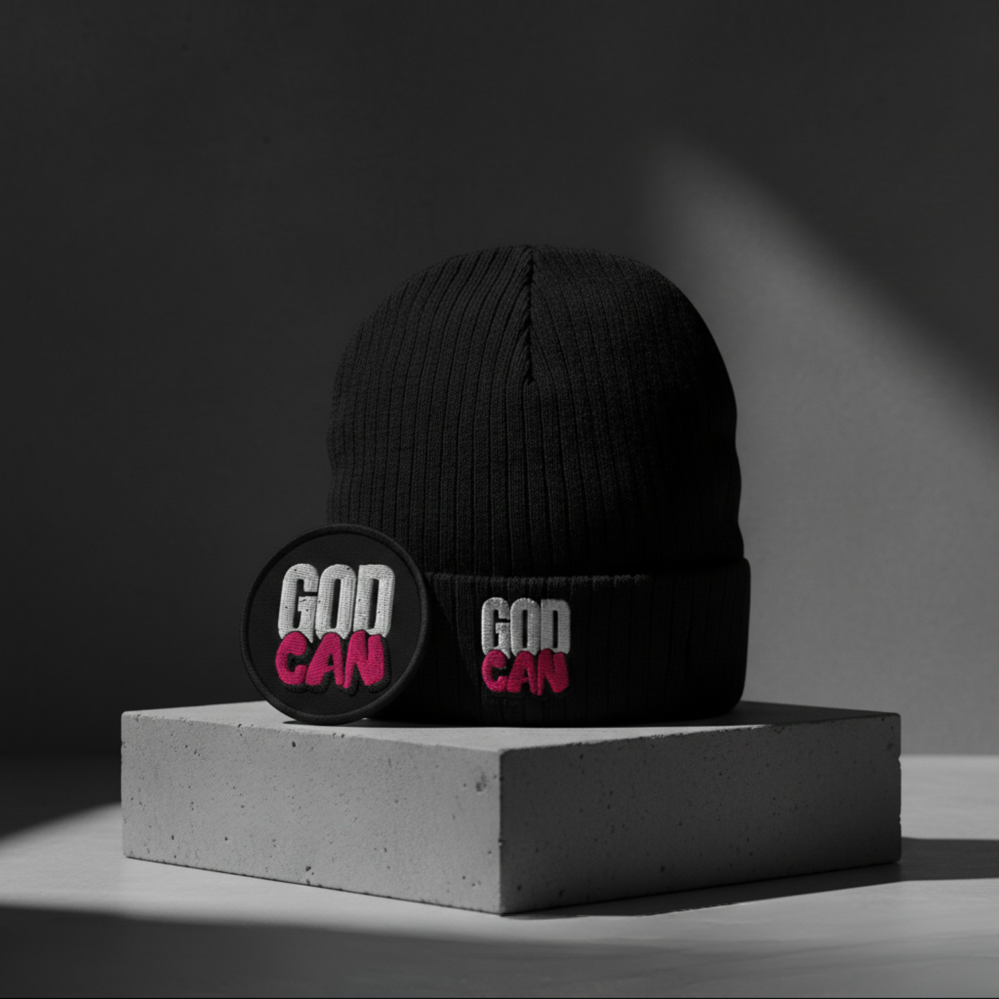 A professional product photograph of the GodCan Foundation Series. A black ribbed beanie with a white and pink embroidered logo sits on a concrete block next to a circular 'GOD CAN' embroidered patch.
