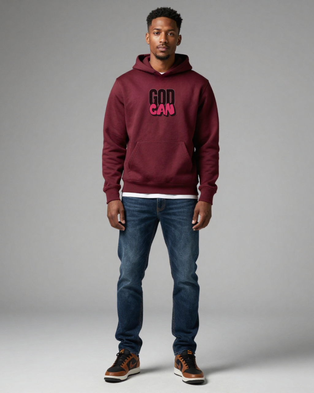 Man wearing a maroon hoodie with text, blue jeans, and brown shoes on a gray background